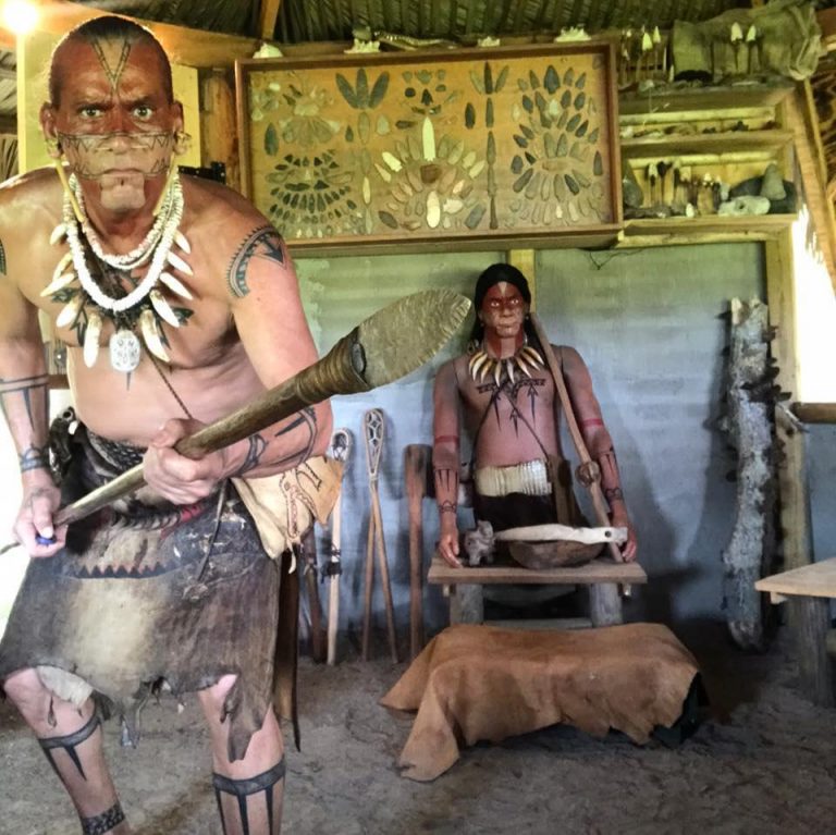 A man holds a spear while a doll sits on a wooden bench. Gonativenow Jororo Village Walkthrough Show Native American Man Spear Doll