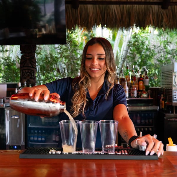 A bartender pours a drink from a shaker into multiple glasses. Junglequeen Ignite The Night Isle Dinner Show With S Bartender Mixing Drinks Bar
