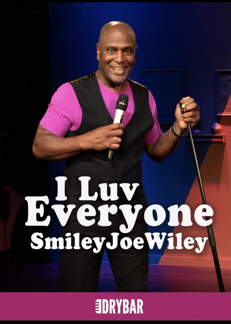 A performer in a pink shirt and black vest holds a microphone on stage. Keeponsmilen Best Comedy Show On Wheels Comedy Show Performer Microphone Stage