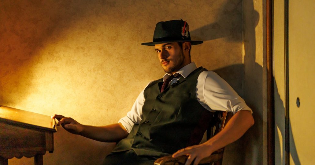 A man in a dark hat and vest sits confidently at a wooden table. Rigatonysclarion Riga Tony S Branson Murder Mystery Dinne Mystery Dinner Show Actor Hat