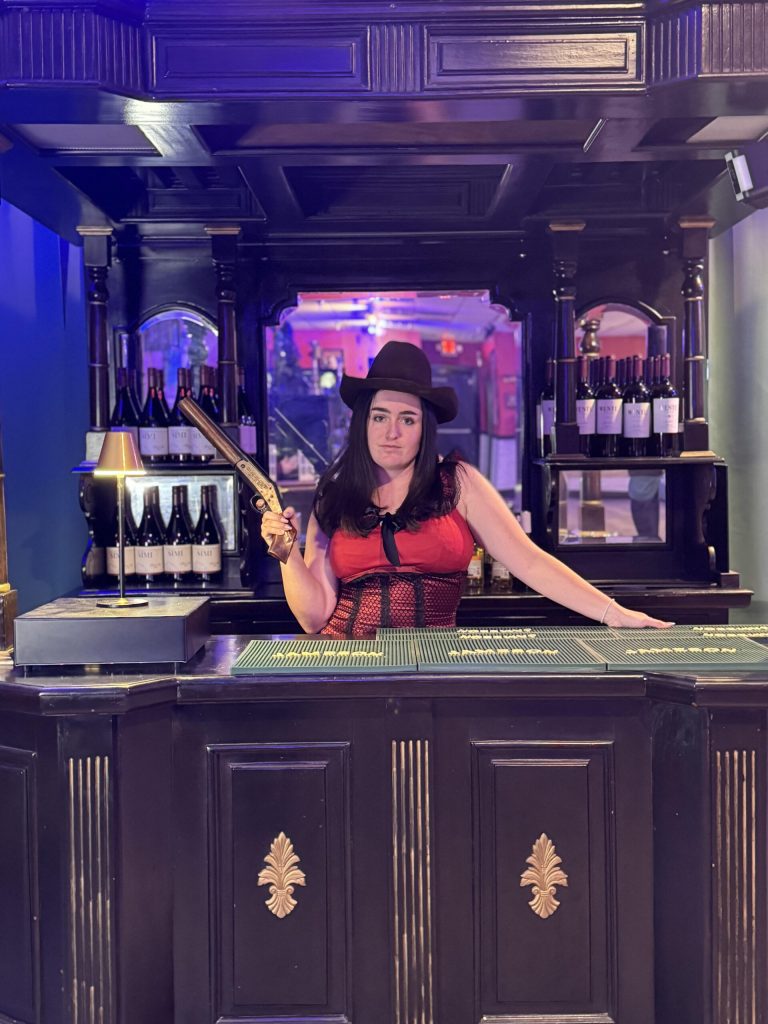 A bartender in a red dress and cowboy hat holds a toy gun at a bar. Rigatonysdinnershow Murder In The Wild West Bartender Western Costume Gun Wine Bottles