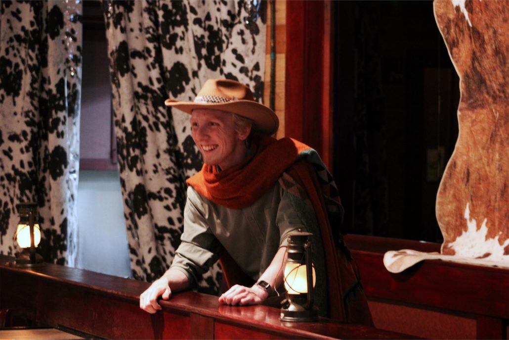 A smiling cowboy actor leans on a wooden bar with a lantern. Rigatonysdinnershow Murder In The Wild West Cowboy Actor Lantern Cowhide