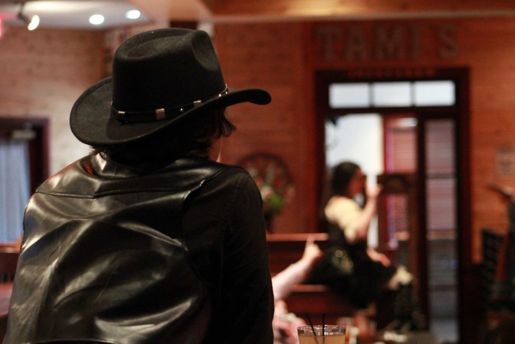A person in a black cowboy hat and leather jacket sits at a table. Rigatonysdinnershow Murder In The Wild West Cowboy Hat Leather Jacket Dinner Show