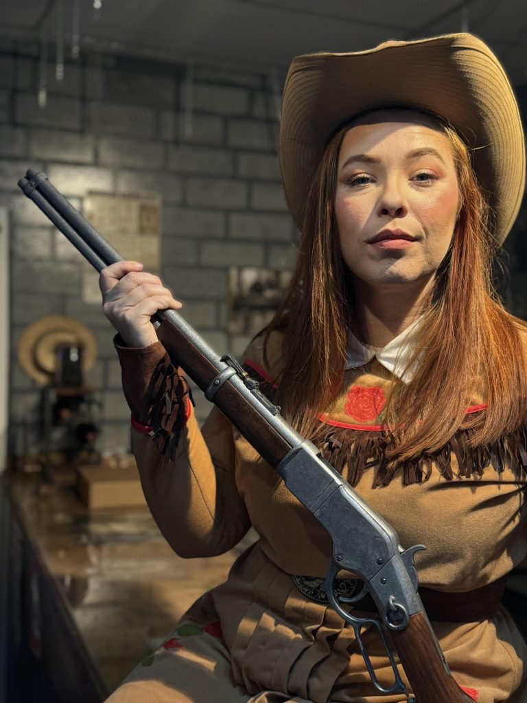 A woman in a cowgirl costume holds a rifle, posing confidently. Rigatonysdinnershow Murder In The Wild West Cowgirl Rifle Costume Dinner Show
