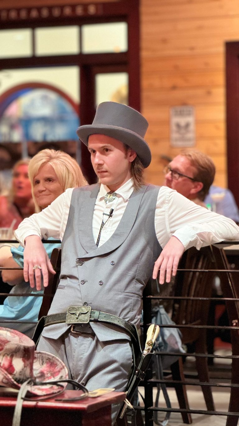 A performer in a gray suit and top hat leans against a railing. Rigatonysdinnershow Murder In The Wild West Dapper Performer Western Dinner Show