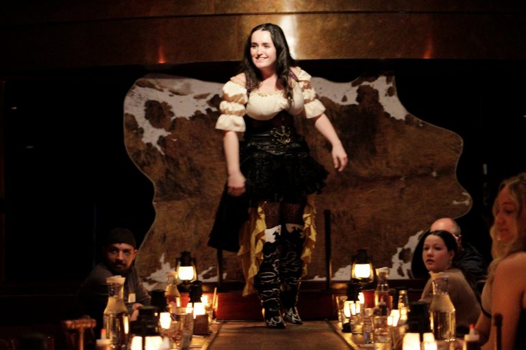 A performer in a western costume walks on a stage with diners watching. Rigatonysdinnershow Murder In The Wild West Dinner Show Performer Western Costume