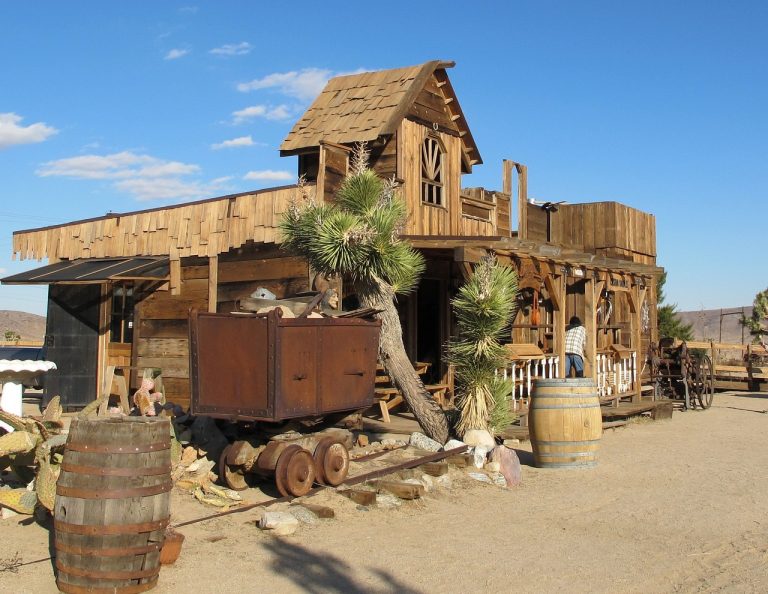 A wooden building with a slanted roof, surrounded by cacti and barrels. Rigatonysdinnershow Murder In The Wild West Wild West Diner Wooden Building Cactus