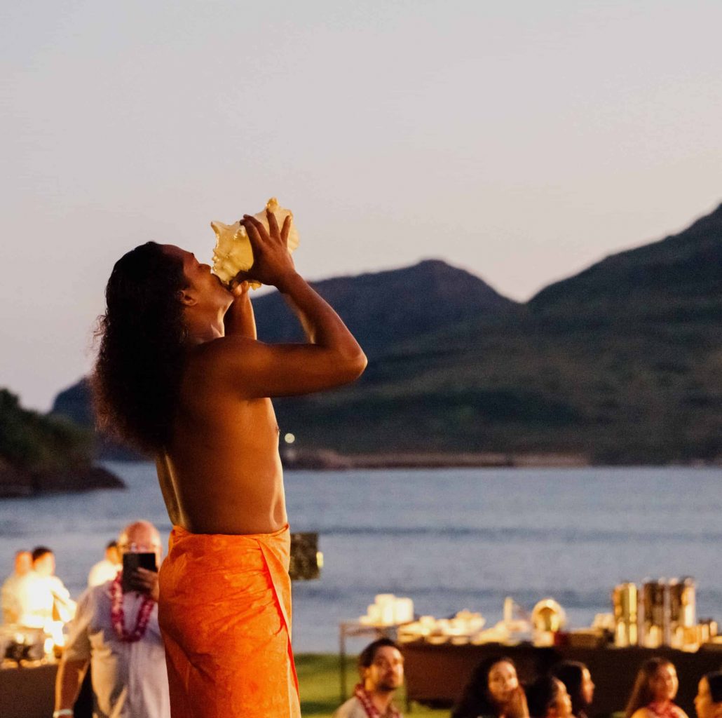 A dancer blows into a large shell with mountains in the background. Royalsonestaluau Drums Dances Of Polynesia At The Royal S Polynesian Dancer Shell Mountains
