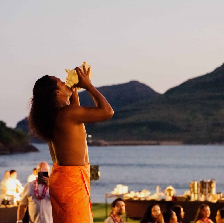 A dancer blows into a large shell with mountains in the background. Royalsonestaluau Drums Dances Of Polynesia At The Royal S Polynesian Dancer Shell Mountains
