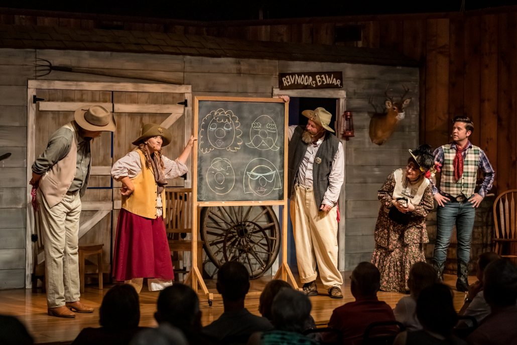 Actors perform in front of a chalkboard displaying cartoon faces. Theshepherdofthehills Branson S Whodunnit Hoedown Murder Myste Murder Mystery Dinner Show Actors