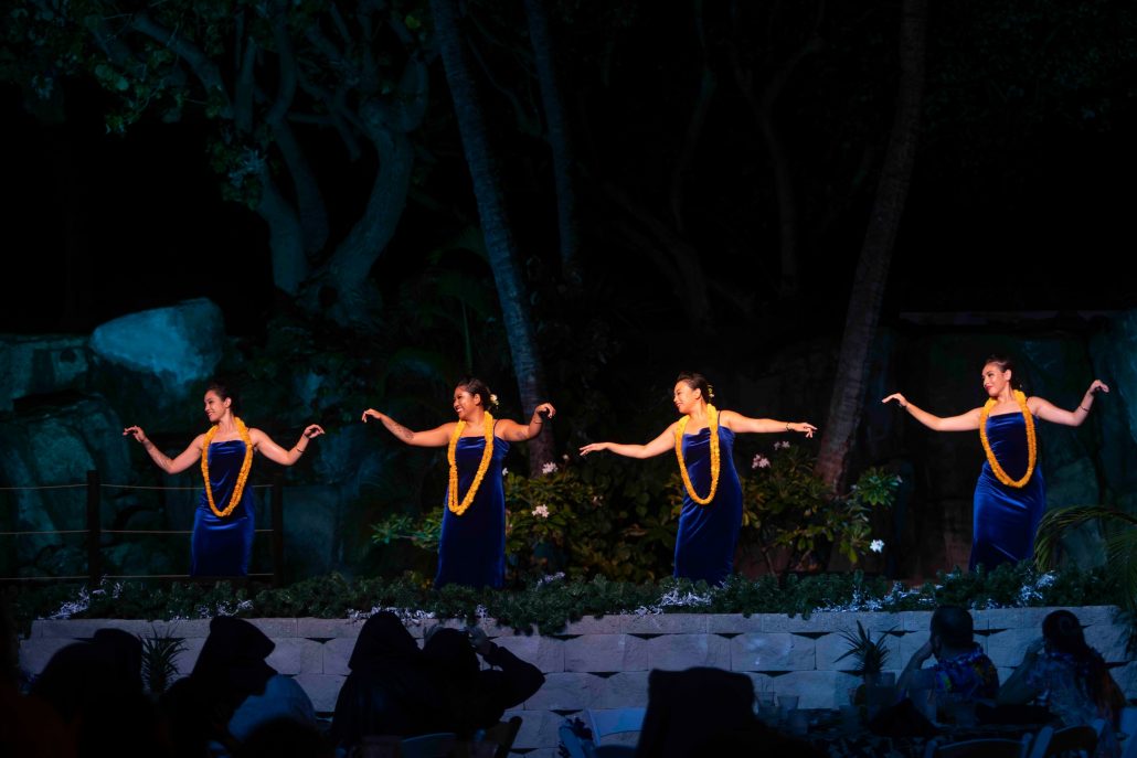 Four dancers in blue dresses and yellow leis perform on stage. Westinmaui Ohana Seating Hula Dancers Blue Dresses Leis