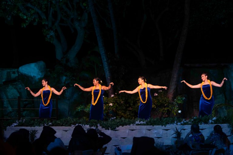 Four dancers in blue dresses and yellow leis perform on stage. Westinmaui Ohana Seating Hula Dancers Blue Dresses Leis