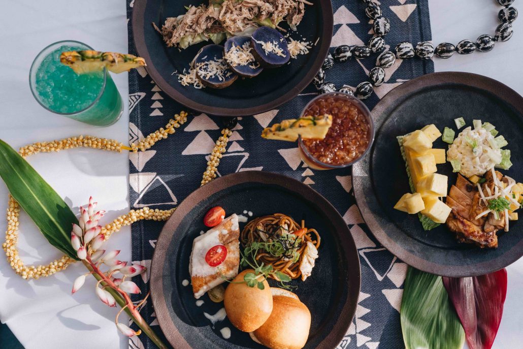 A colorful spread of luau dishes and drinks on a patterned tablecloth. Westinmaui Ohana Seating Luau Food Drinks Tropical Decor