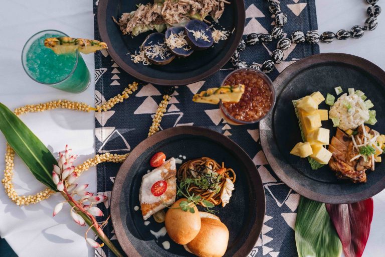 A colorful spread of luau dishes and drinks on a patterned tablecloth. Westinmaui Ohana Seating Luau Food Drinks Tropical Decor
