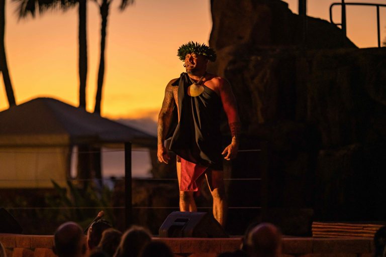 A performer in traditional attire stands on stage during sunset. Westinmaui Ohana Seating Performer Luau Sunset Stage