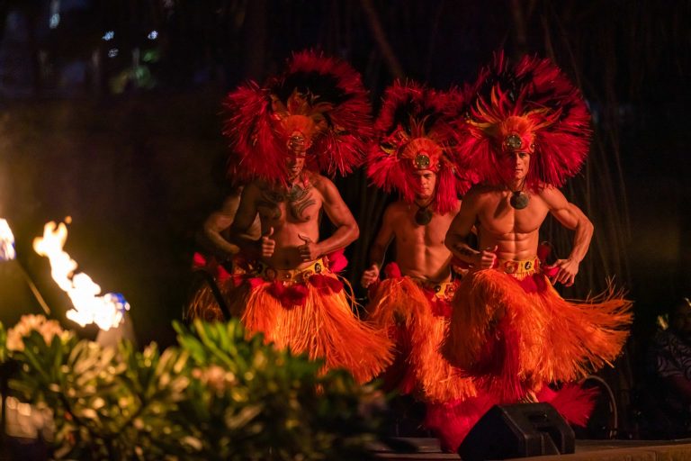 Three male performers in vibrant feathered costumes dance near flames. Westinmaui Ohana Seating Performers Fire Dance Costumes