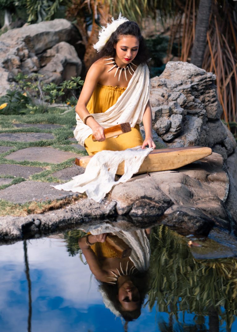 A woman in a yellow dress sits by a pond, holding a wooden tool. Westinmaui Ohana Seating Woman Yellow Dress Luau Reflection
